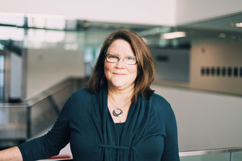 Melody Petlock leans on a railing in the Purdy Crawford Centre for the Arts. She is smiling and wearing glasses, a silver necklace, and a blue cardigan