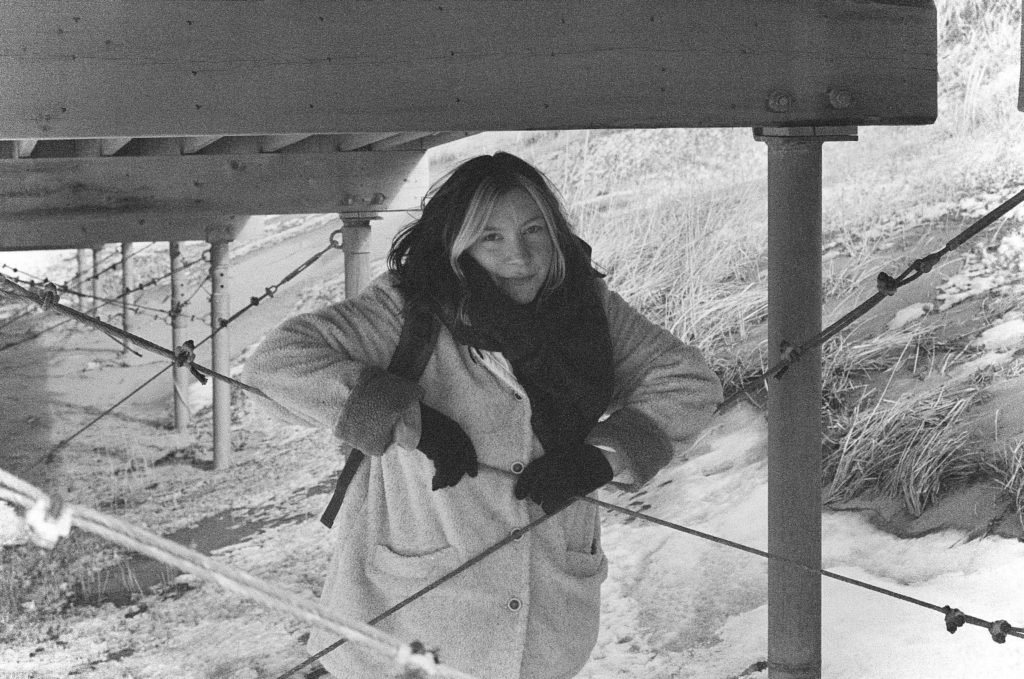 Emma Delaney leans on wire stabilizers under a wooden structure. She is wearing gloves, a scarf, and a big white coat. There is snow on the ground in the background. There is sand and grass. The photo is black and white and shot on film.