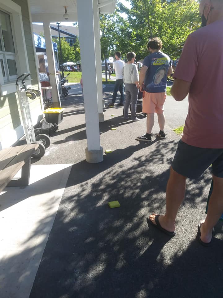 Market-goers stand in line behind a booth. They are standing by social distance markers. There are 5 people in line. It is sunny outside.