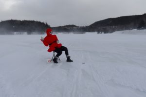 The Queer Mummer is dressed in winter clothes and sits on a folding chair in the middle of a frozen body of water.
