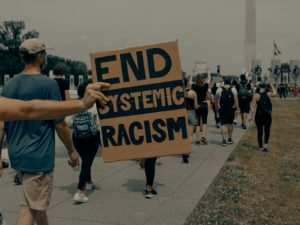 A hand holds a cardboard sign that says "End system racism." People walk in the background.