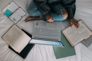 A person sits cross-legged on a bed an looks at a laptop. The picture is bird's eye view, and there are binders and books open and scattered around the bed.