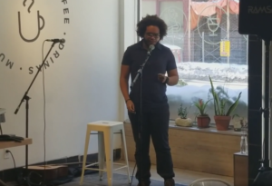 Thandiwe McCarthy stands inside of a cafe in front of a microphone and speaks.